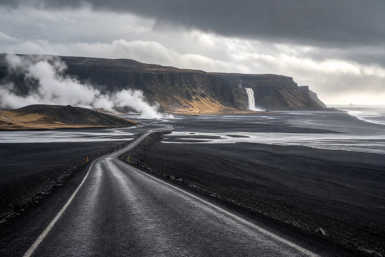 An Icelandic road crosses black sand toward geothermal steam and a distant waterfall.
